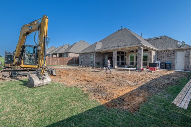 Excavator grading a backyard next to a suburban home during foundation prep and landscaping construction